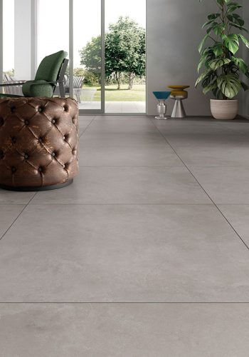Ceramica Valsecchia Recover 'Grey', lichtgrijze betonlook vloertegels bij een glazen pui met een leren poef en tuinuitzicht