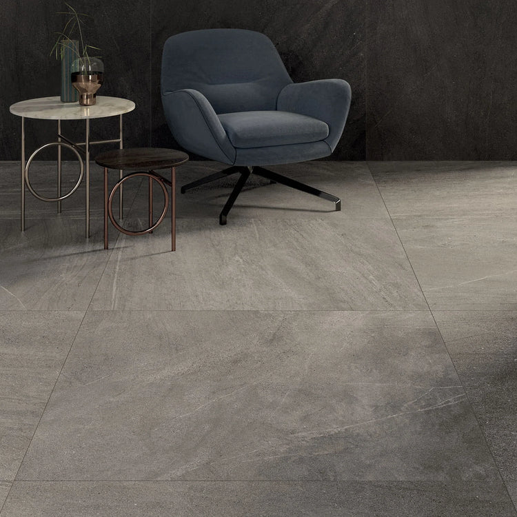 Lea Ceramiche Nextone, grijze vloertegels met natuursteenlook bij een donkere wand, met blauwe fauteuil en ronde bijzettafels