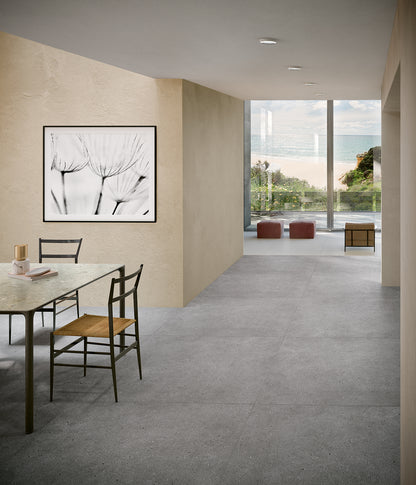 EnergieKer Brera 'Glow', grijze betonlook vloertegels in een eetkamer die doorloopt naar een zithoek met zeezicht