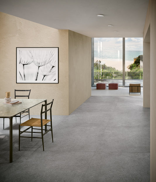 EnergieKer Brera 'Glow', grijze betonlook vloertegels in een eetkamer die doorloopt naar een zithoek met zeezicht